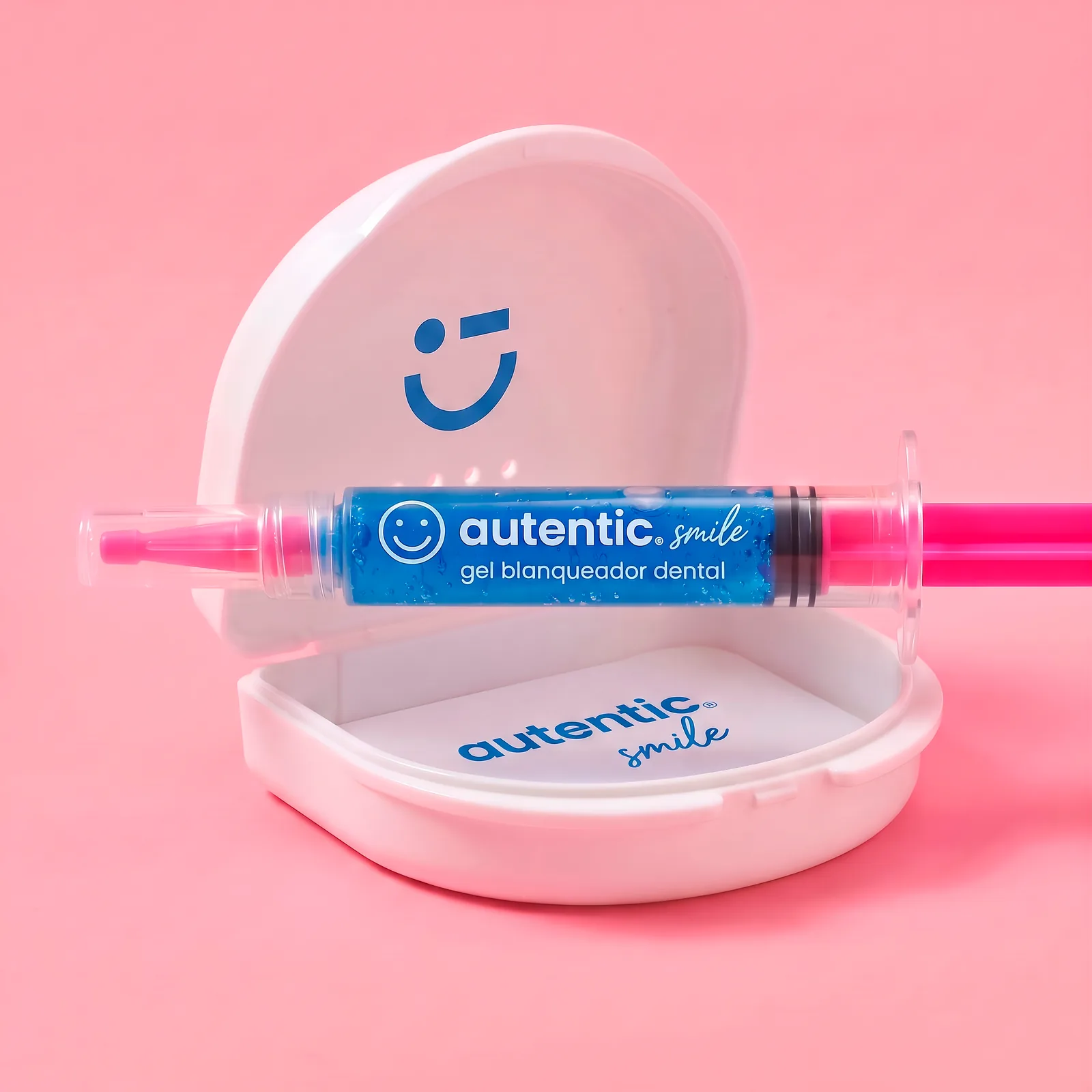 Gel blanqueador dental Autentic Smile en jeringa junto a su estuche, sobre fondo rosa